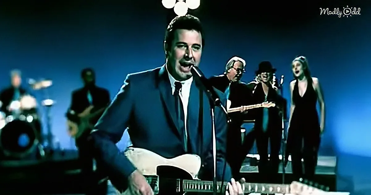 A man in a suit sings into a microphone while playing guitar on stage with a band.