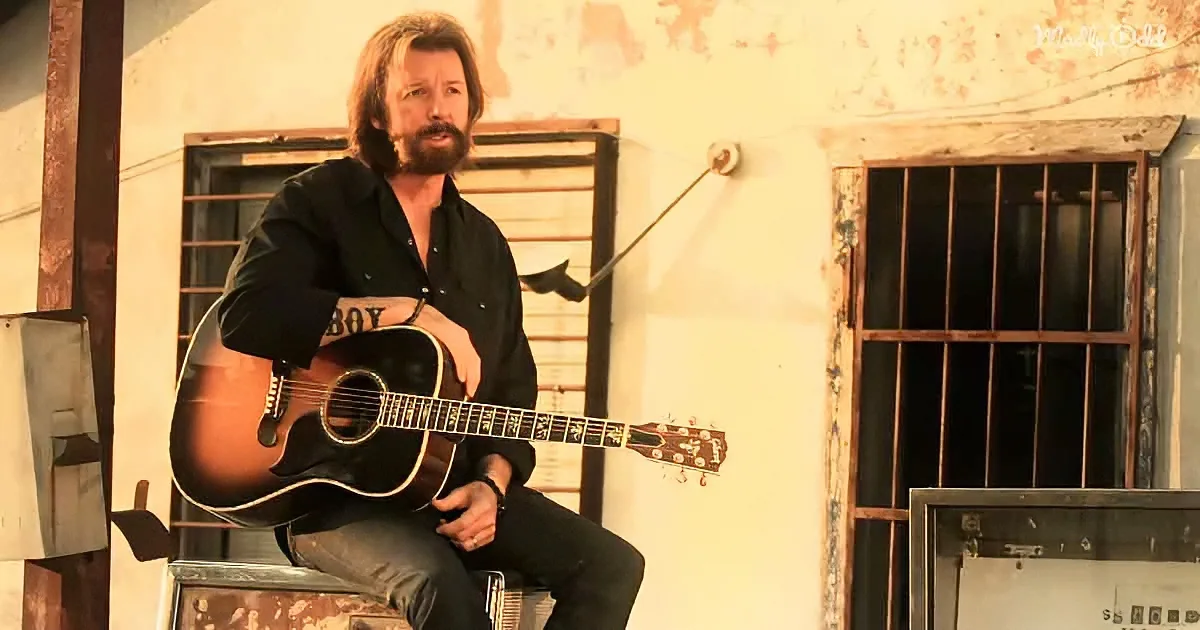 A man with a beard sits on a metal box, holding an acoustic guitar, in front of a weathered building with barred windows.
