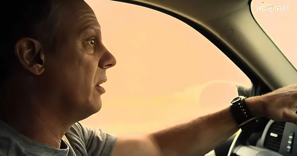 A man drives a car, wearing a watch, with a focused expression and a warm, orange-tinted background.