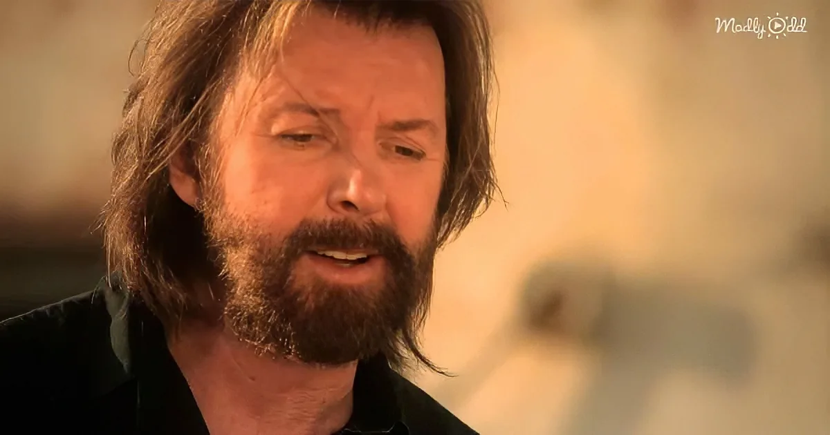 A man with long hair and a beard, wearing a black shirt, appears to be speaking or singing.