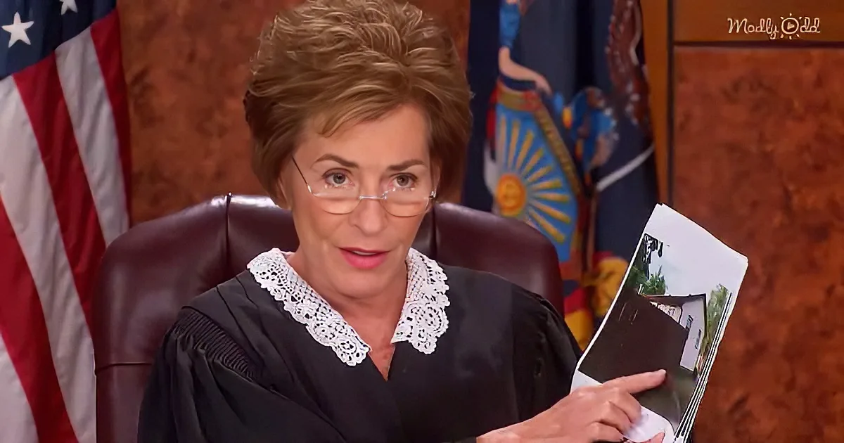 A woman in a judge's robe holds a photo, seated in a courtroom with flags behind her.