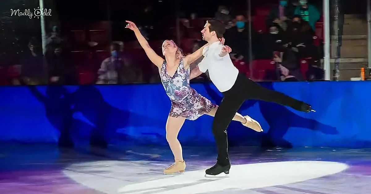 A figure skating couple performs a lift on ice, with the woman in a floral dress and the man in a white shirt and black...