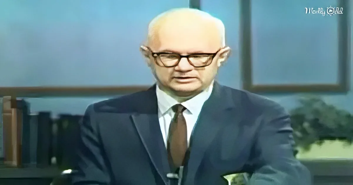 A man in a suit and glasses sits at a desk, looking downward, with a blurred background and a "Madly Odd" watermark.