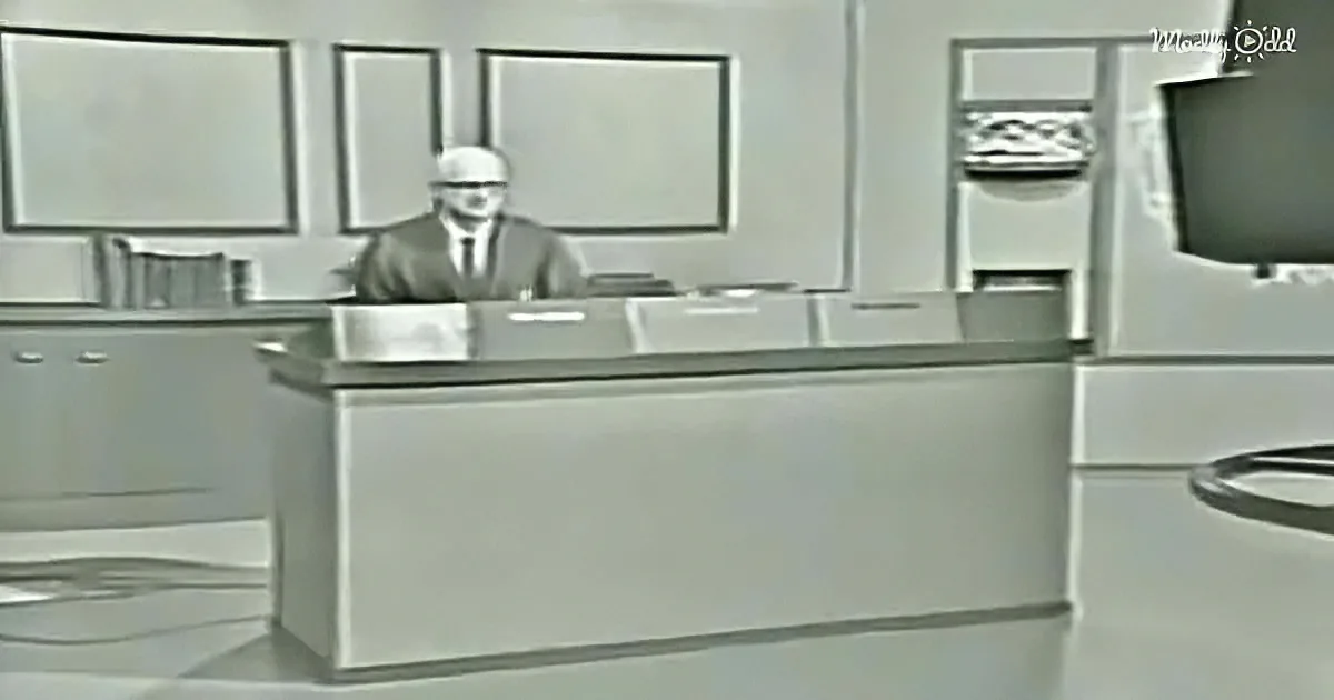 A man in a suit sits behind a large, futuristic desk in a minimalist, monochromatic room.