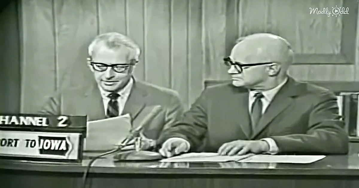 Two men in suits sit at a desk with microphones, one reading from a paper, in a studio setting.