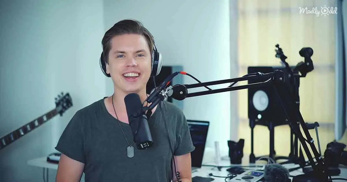 A smiling person wearing headphones stands in front of a microphone in a room with musical equipment.