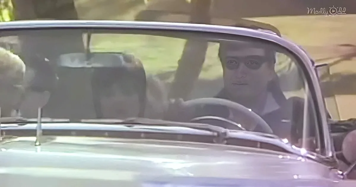 A man wearing sunglasses sits in the driver's seat of a classic convertible car.