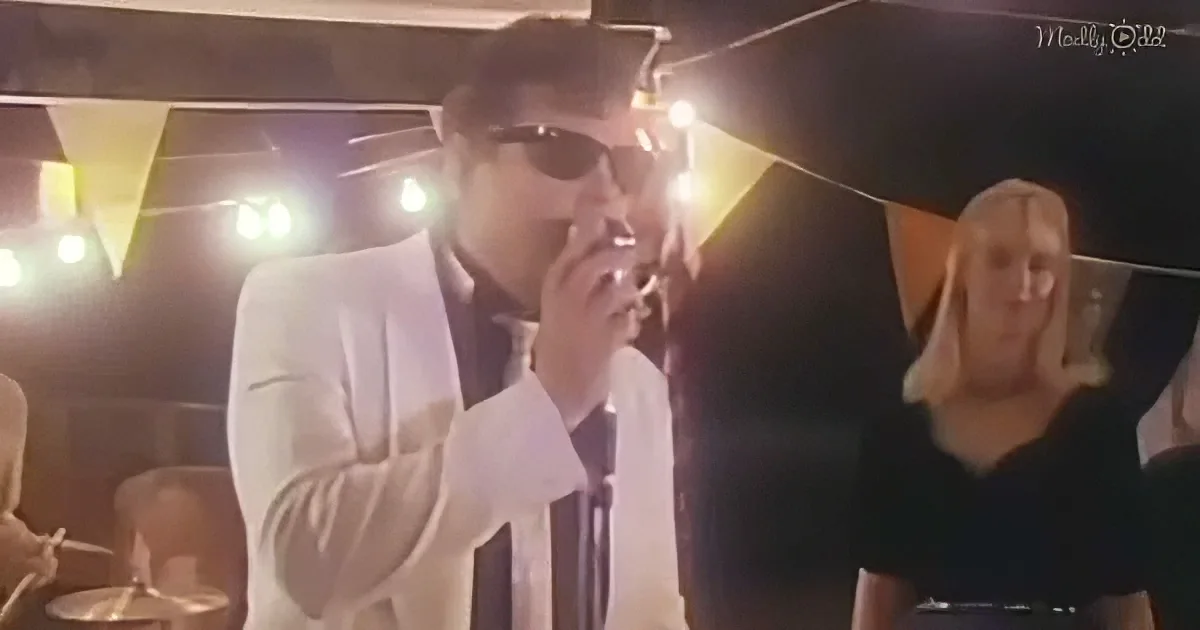 A man in a white suit sings into a microphone, illuminated by stage lights, with a woman in the background.