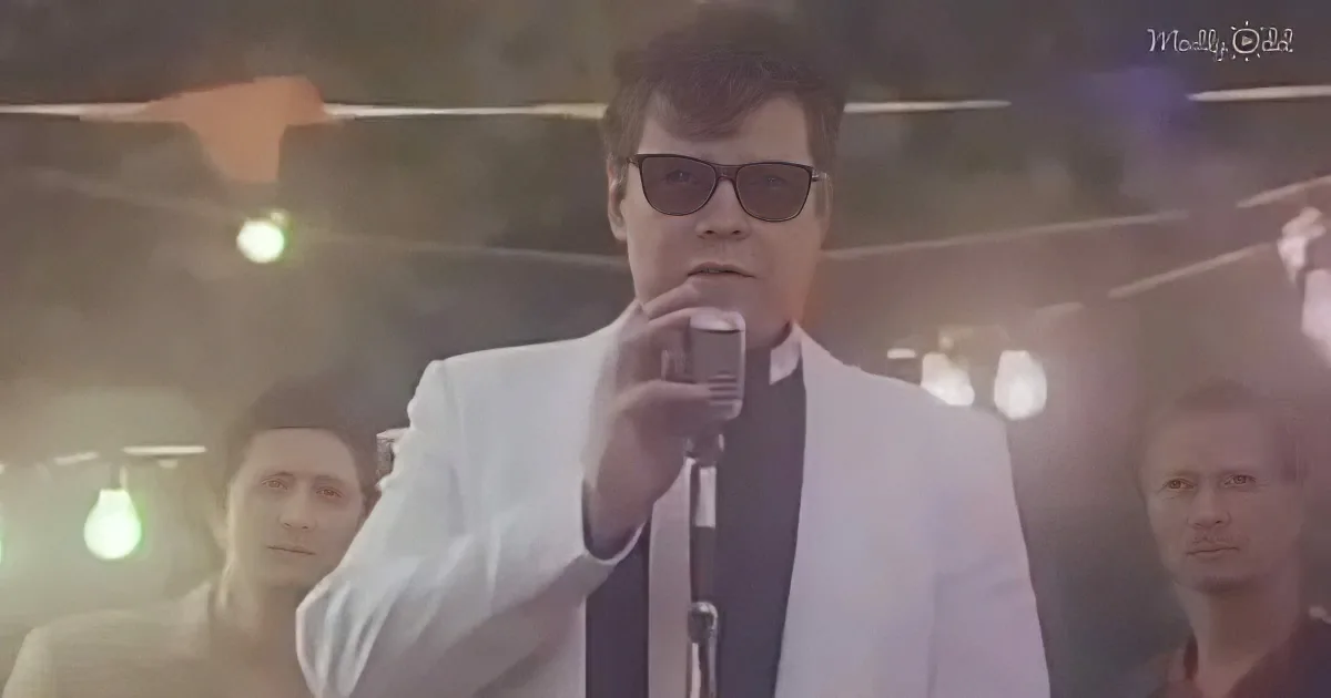 A man in a white suit and sunglasses sings into a microphone on stage, with other musicians in the background.
