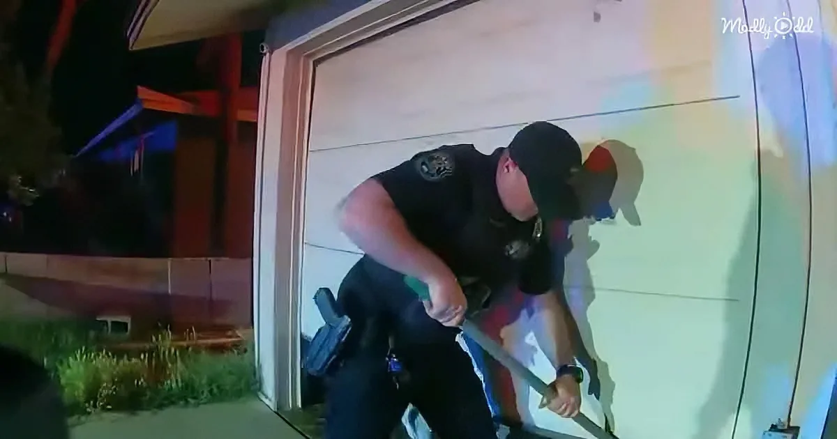 A police officer in uniform is crouching near a garage door, holding a tool.