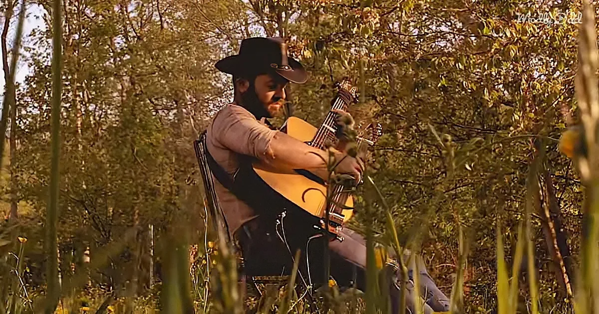 A man in a cowboy hat plays an acoustic guitar in a forested area with tall grass and trees.
