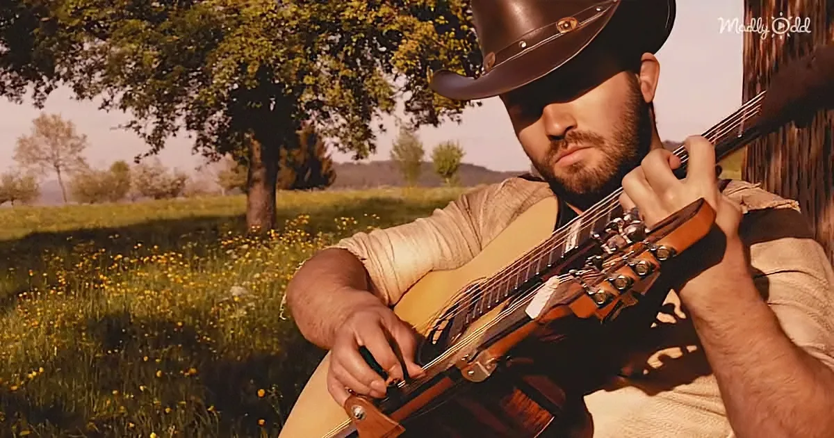 A man in a cowboy hat plays a guitar in a sunlit field with trees and wildflowers.