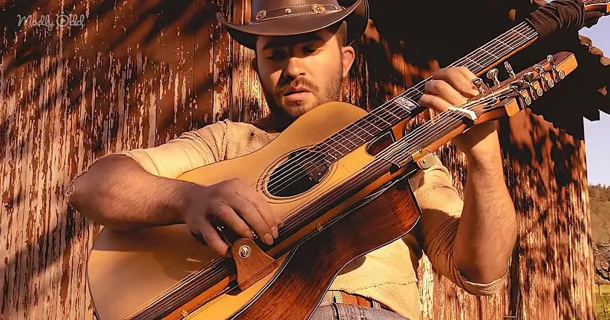 A man in a cowboy hat plays an acoustic guitar against a rustic wooden wall.