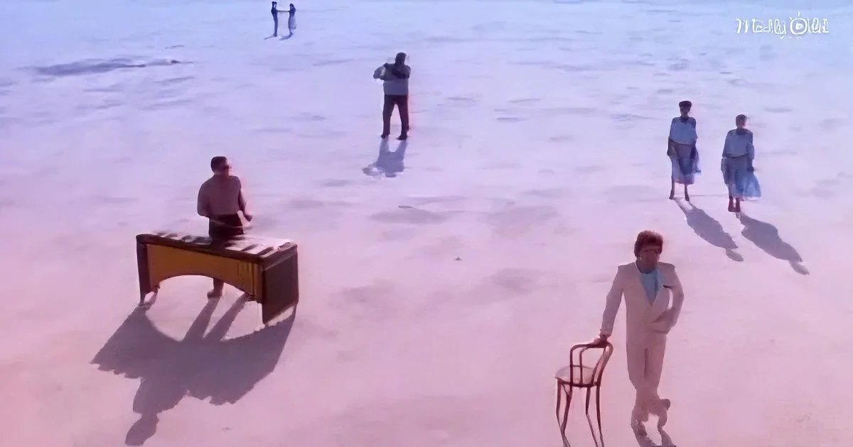 A man in a white suit stands with a chair, while another plays a xylophone on a beach with people in the background.
