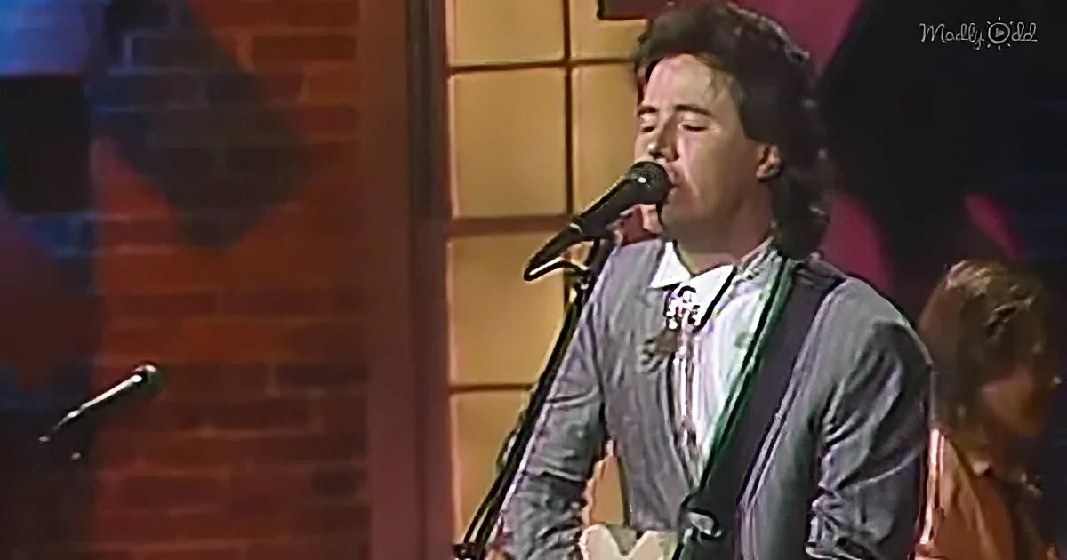 A man in a gray suit sings into a microphone on stage, with a brick wall and another person partially visible.