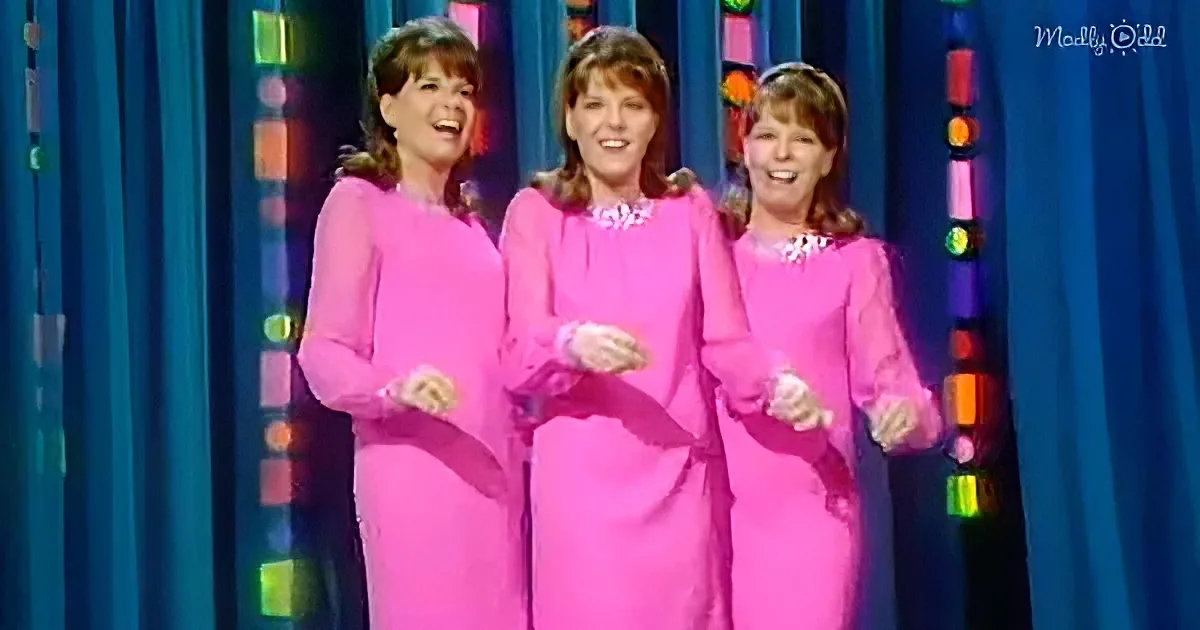 Three women in matching pink dresses stand together, smiling and clapping, against a colorful backdrop.