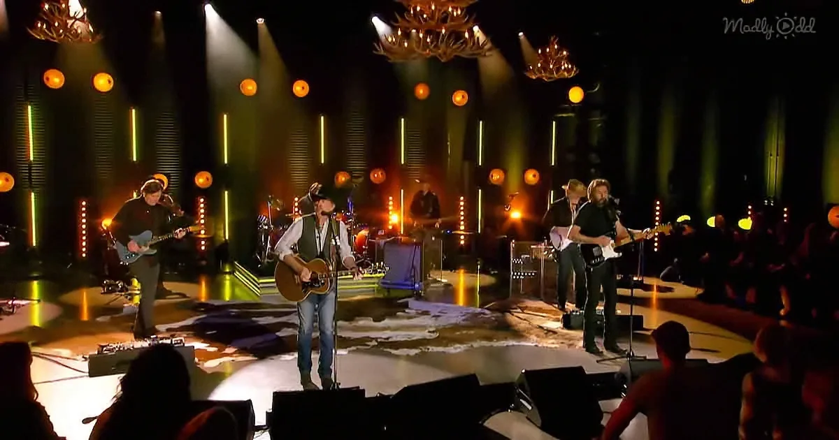 A man in a cowboy hat sings on stage with a band, surrounded by bright lights and chandeliers.