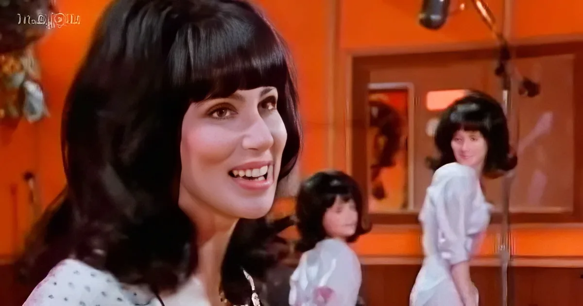 A smiling woman with dark, shoulder-length hair stands in front of a mirror, with two other women in white shirts behind her.