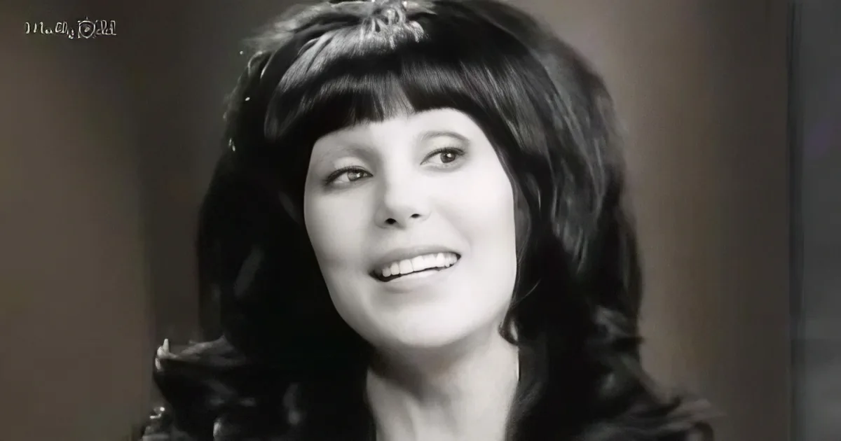 A smiling woman with dark, wavy hair and bangs, wearing a dark outfit, against a blurred background.