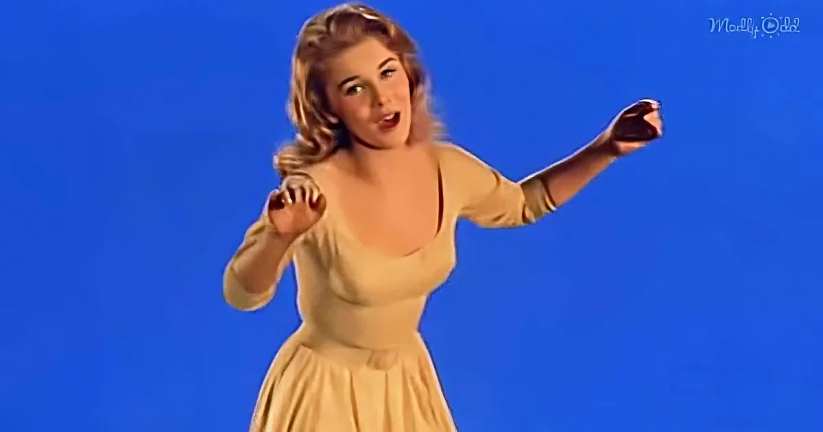 A woman in a beige dress sings with raised arms against a blue background.