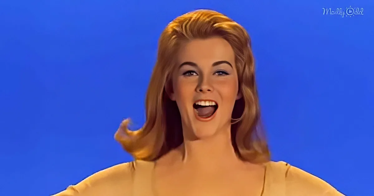 A woman with blonde hair in a light-colored top smiles widely against a blue background.