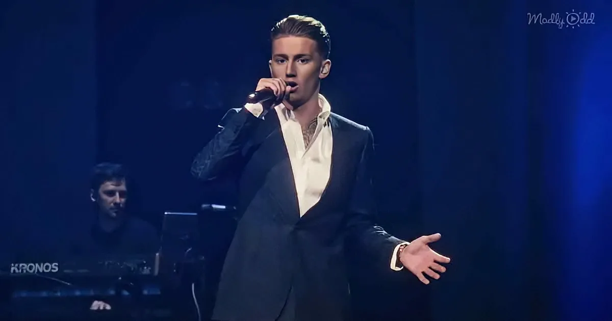 A man in a suit sings into a microphone on stage, with a keyboard player in the background.