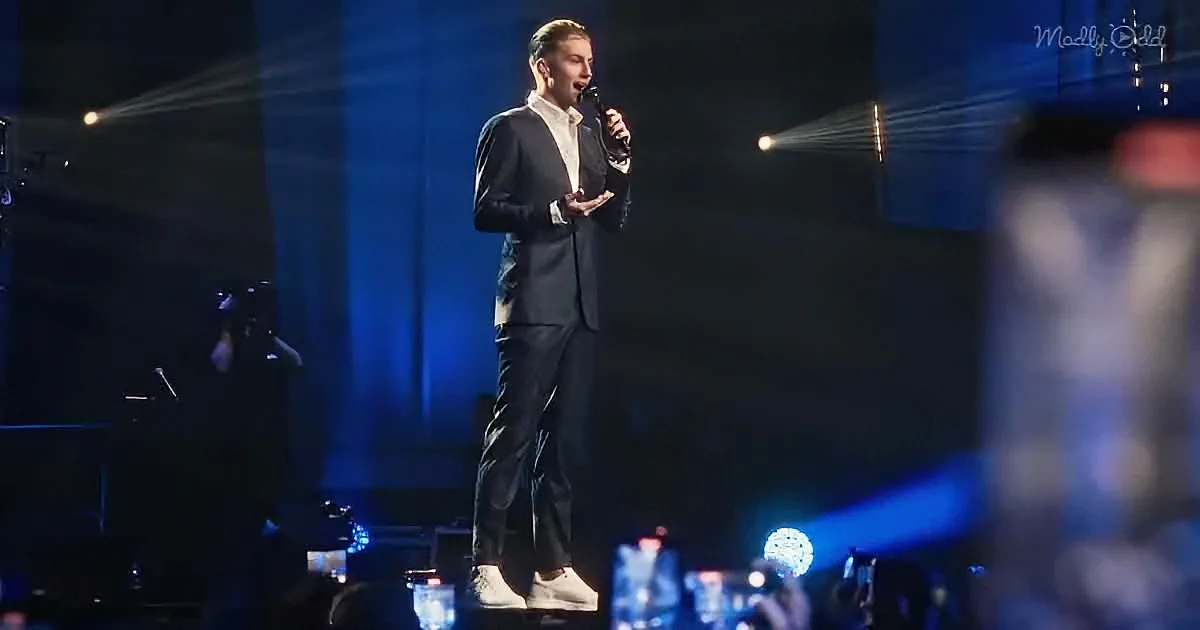 A man in a suit stands on stage holding a microphone, illuminated by stage lights.