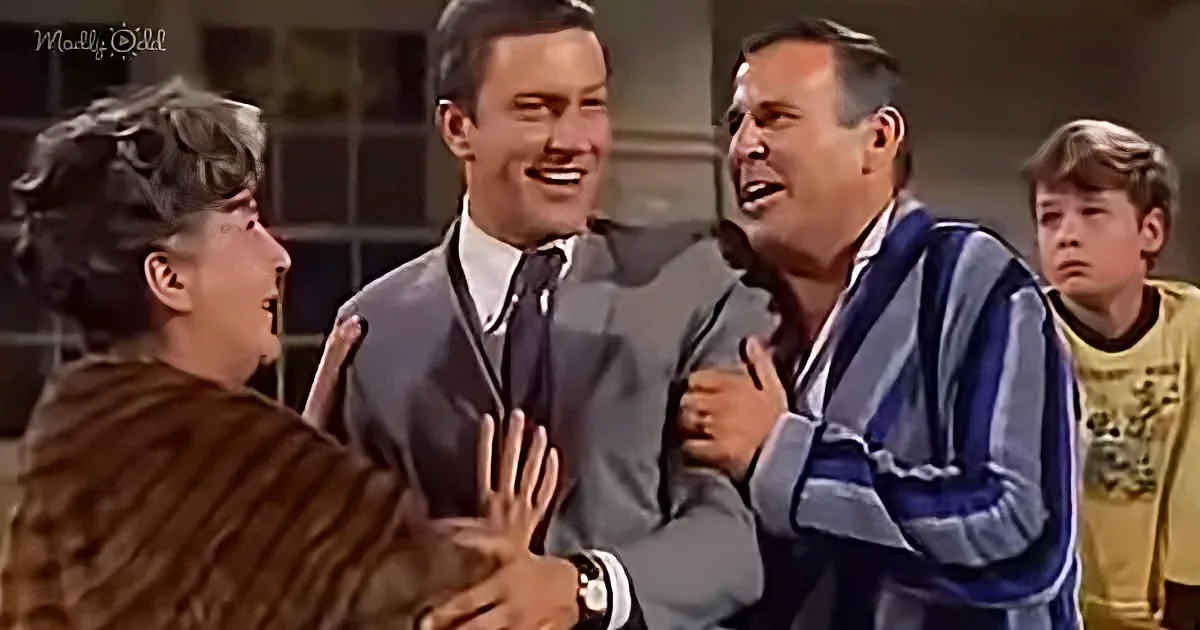 A man in a striped shirt is being embraced by another man in a suit, while a woman and a boy look on.