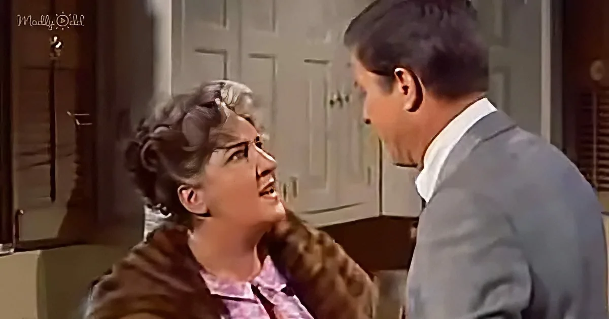 A woman in a fur coat and pink blouse is talking to a man in a suit in a kitchen setting.