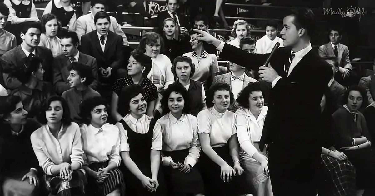 A man in a suit points at a seated audience of students in a gymnasium.