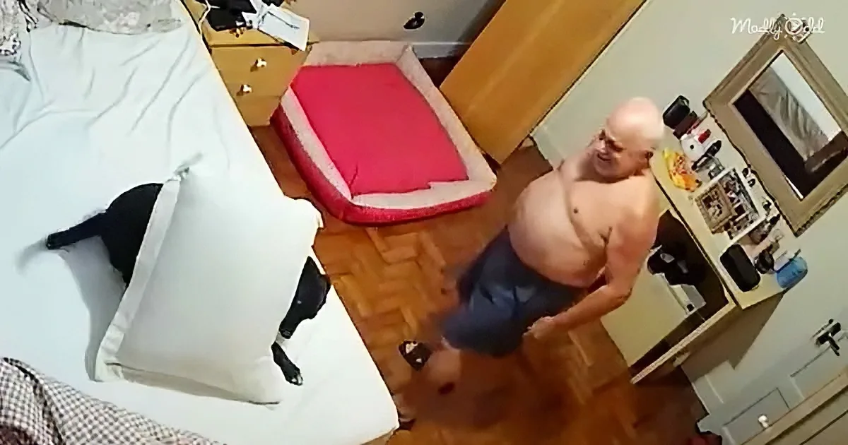 A shirtless man in blue shorts stands in a room with a bed, dresser, and mirror.