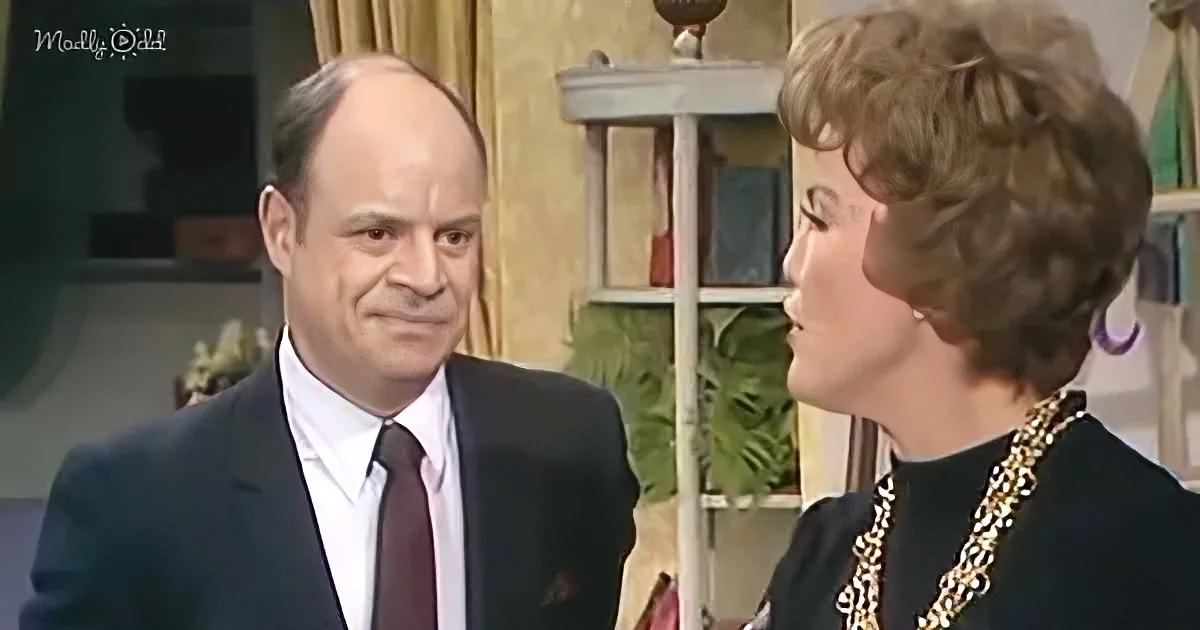 A man in a suit and tie is looking at a woman with short hair, both in a room with plants and shelves.