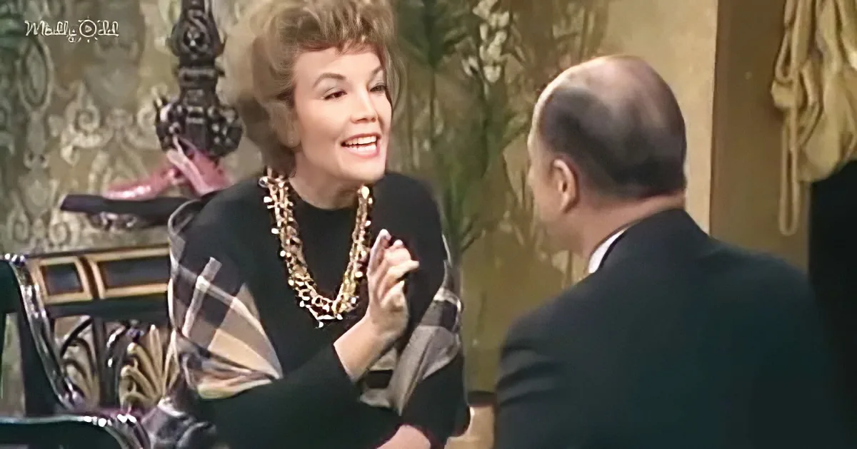 Woman in black dress with gold necklace gestures while seated, smiling at man in suit.