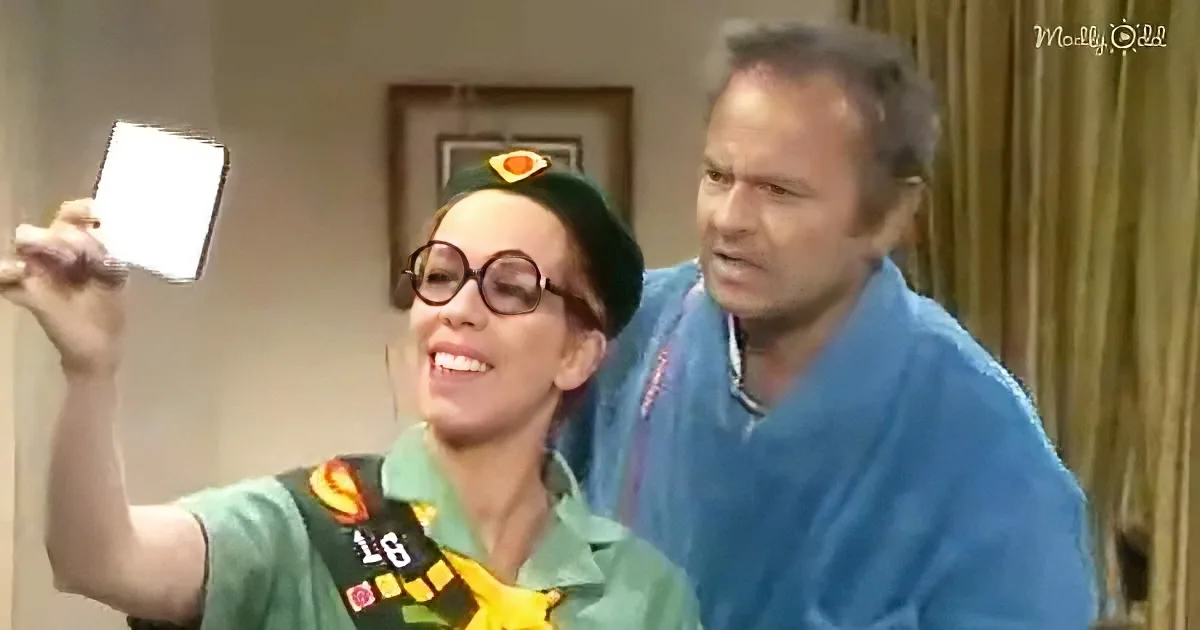 A woman in a green uniform takes a selfie with a man in a blue shirt behind her.