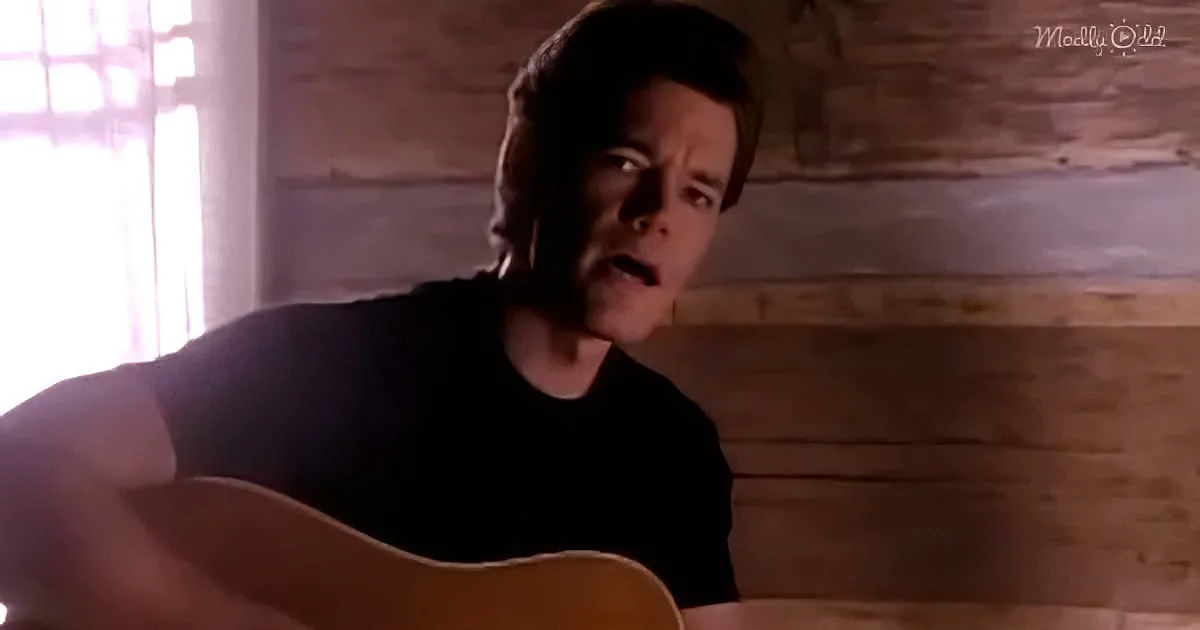 A man in a black shirt plays an acoustic guitar in a rustic setting with wooden walls.