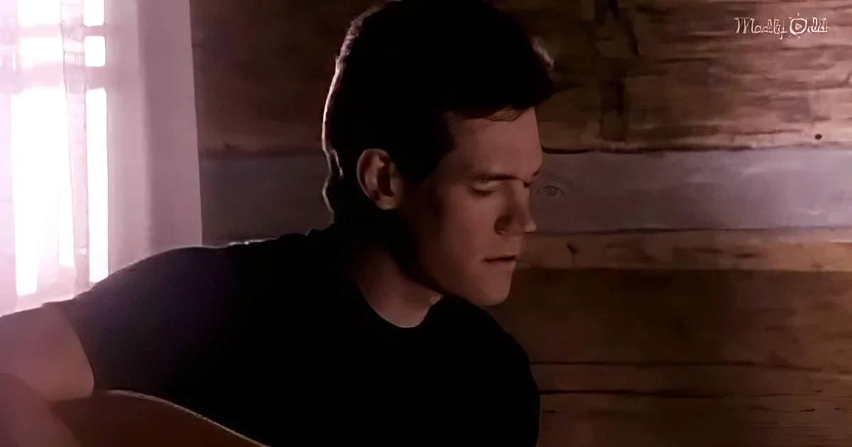 A man in a black shirt plays a guitar in a dimly lit room with wooden walls.