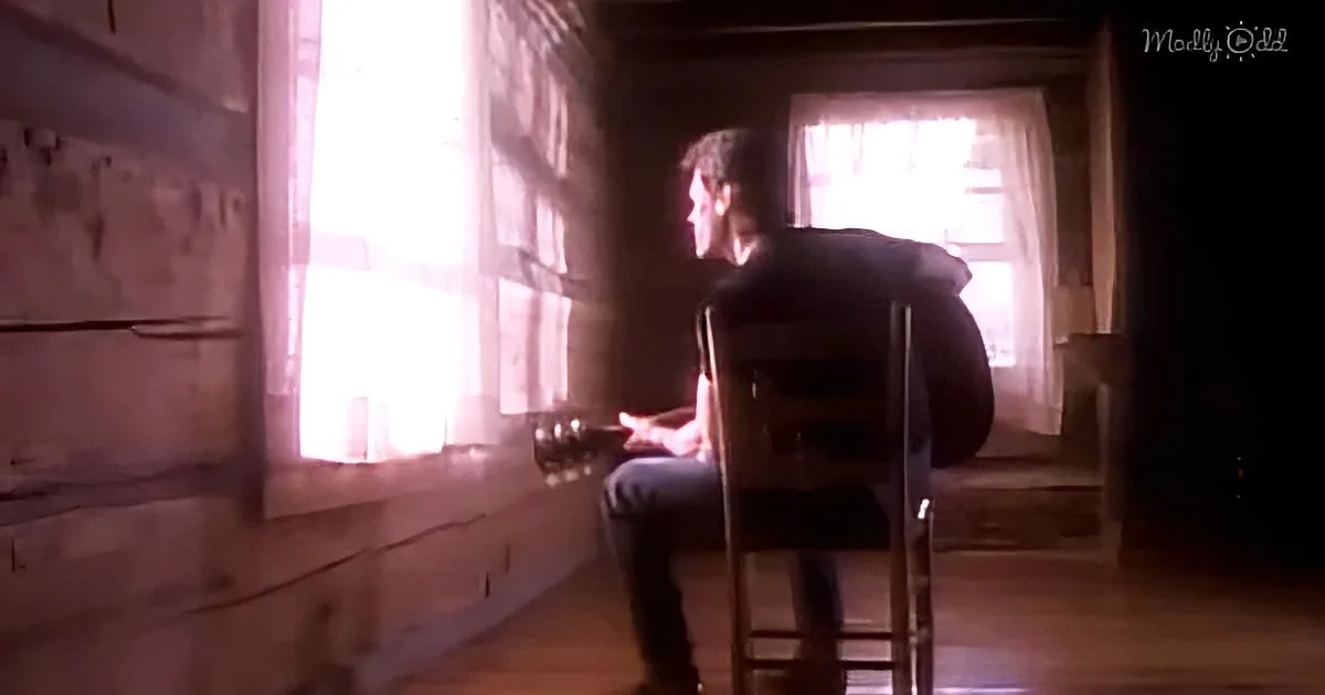 A man sits on a chair, gazing out of a window with light streaming in, in a dimly lit room.