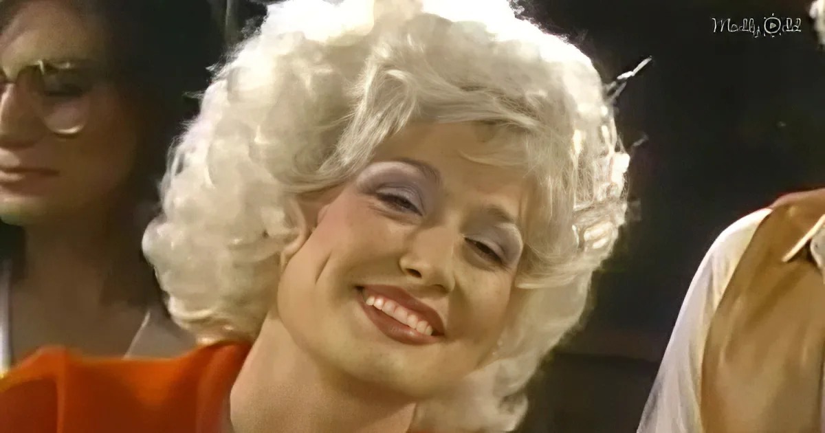 A person with white curly hair and makeup smiles, wearing an orange outfit, with others in the background.