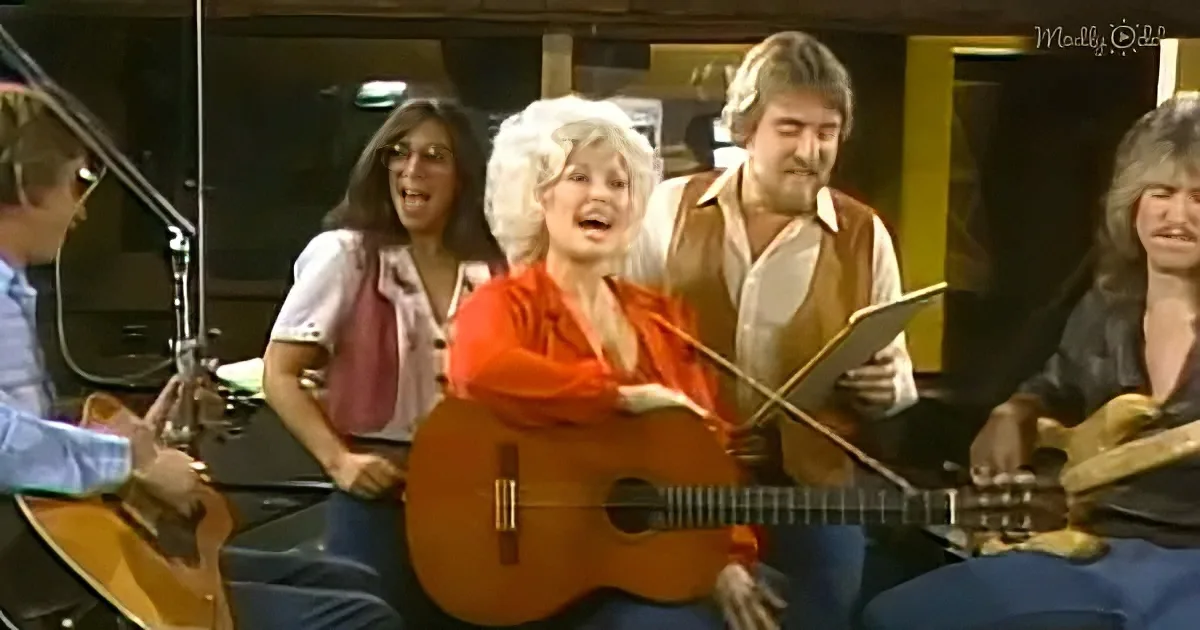 A woman in a red shirt plays guitar, surrounded by musicians in a recording studio.
