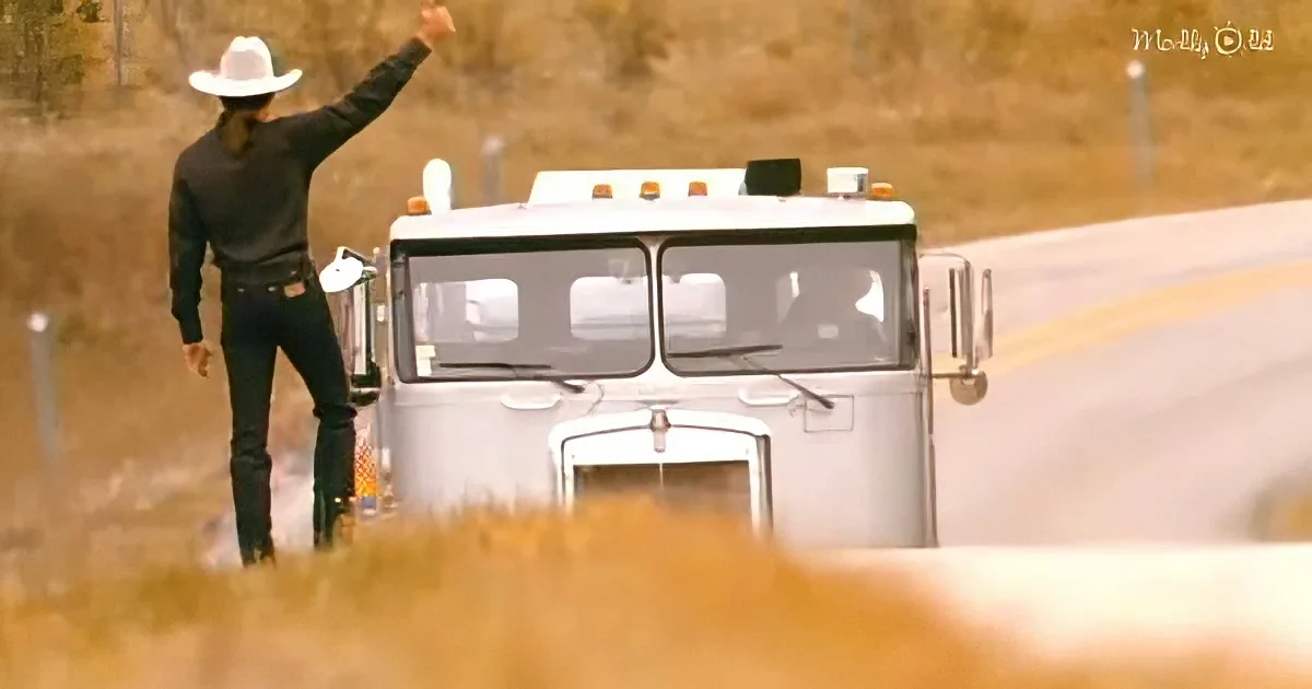 A man in a cowboy hat and black attire stands beside a large truck, raising his arm.