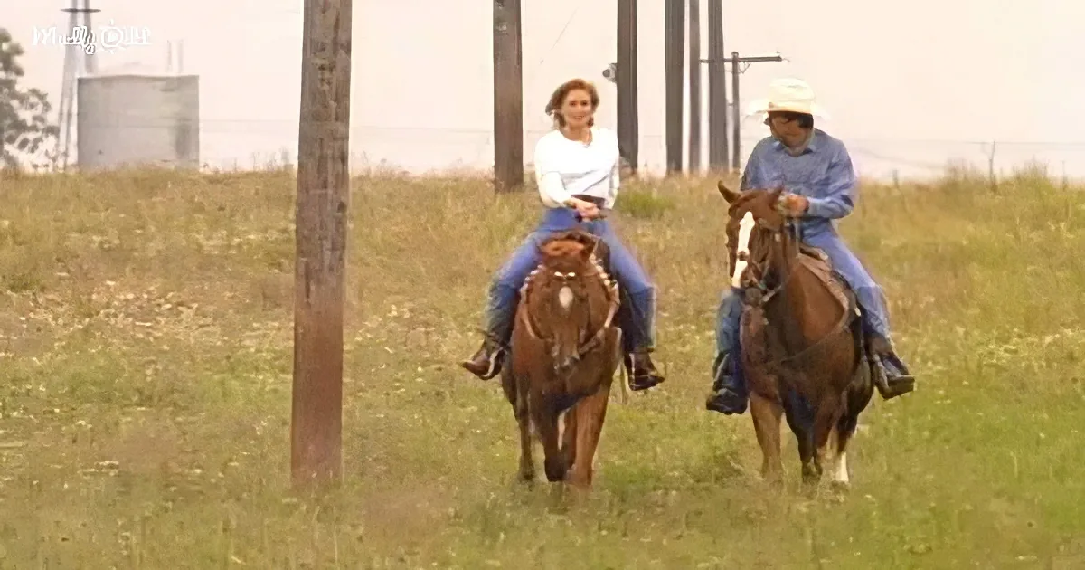 Two people on horses in a grassy field, one wearing a cowboy hat.