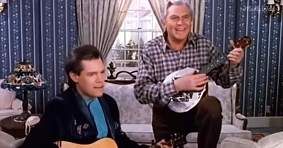A man in a plaid shirt holds a guitar, smiling, while another man in a black jacket sits, holding a guitar, in a cozy room.