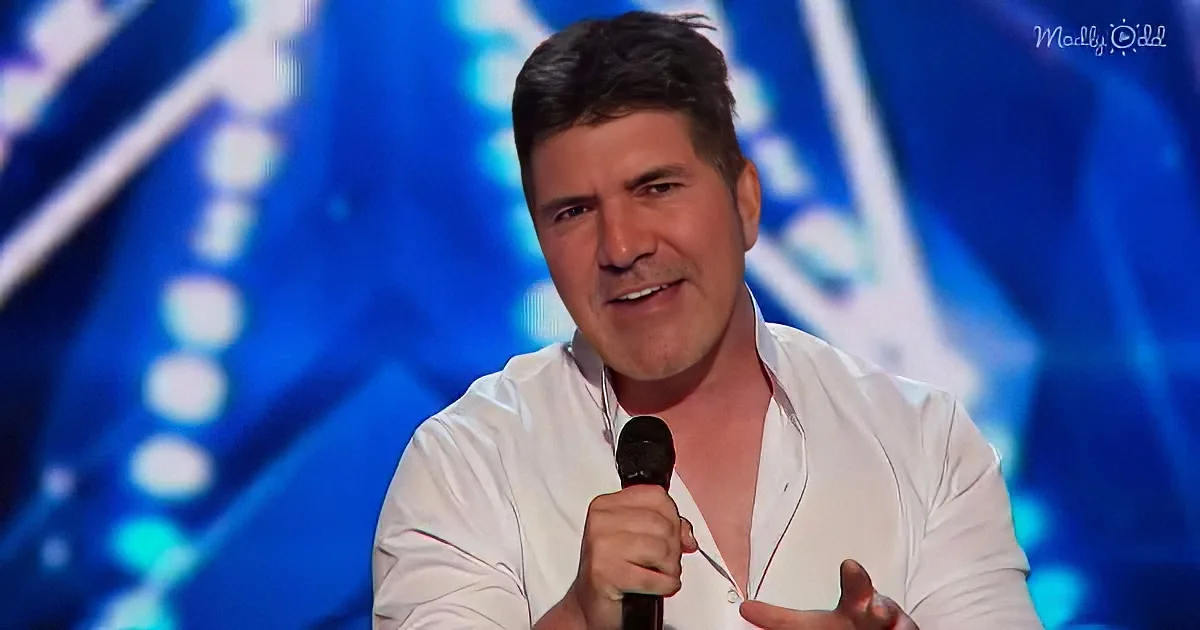 A man in a white shirt speaks into a microphone, holding a card, against a blue, illuminated background.