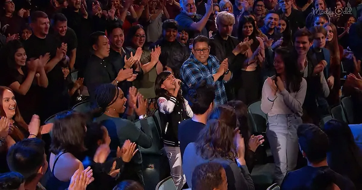 A woman in a black jacket and white shirt is singing on stage, surrounded by an audience clapping and cheering.