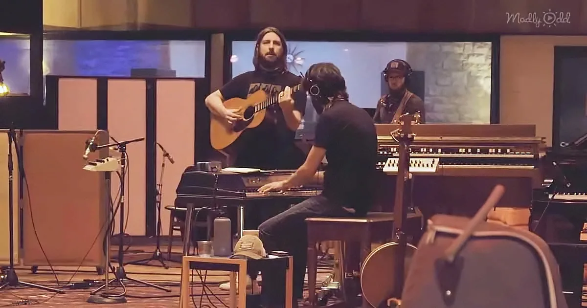 A man plays guitar in a recording studio with another man at a keyboard and a third person in the background.