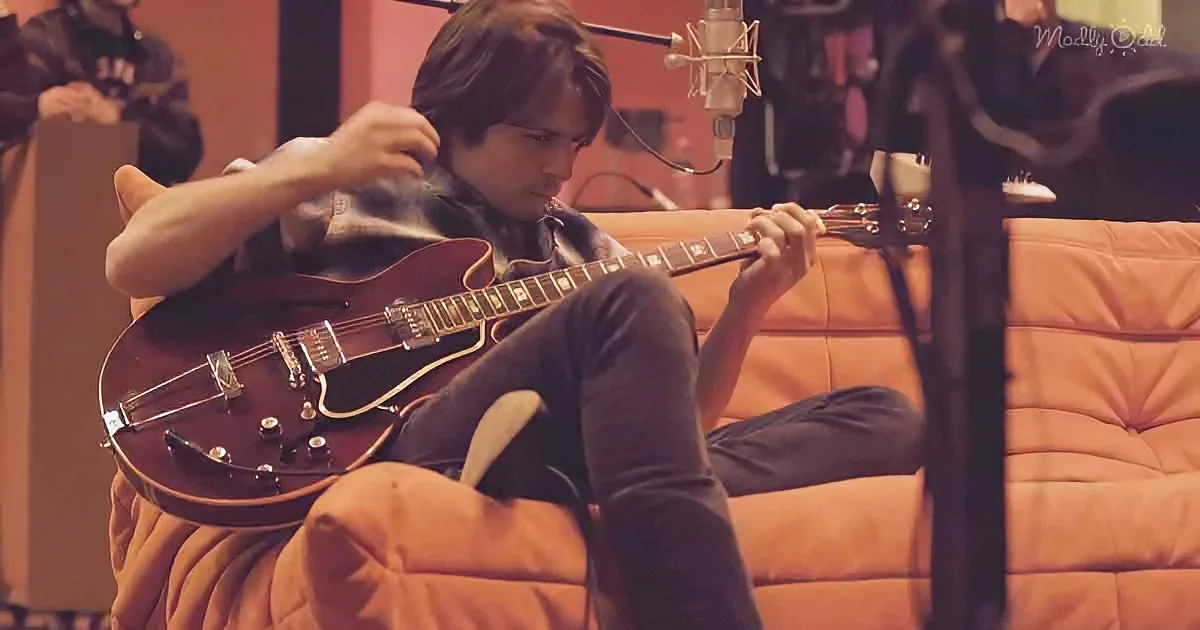 A person sits on a couch, playing a dark brown electric guitar in a recording studio setting.