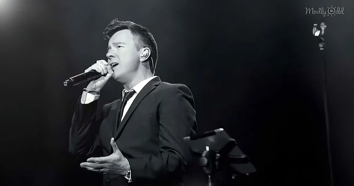 A man in a suit sings into a microphone on stage, illuminated against a dark background.