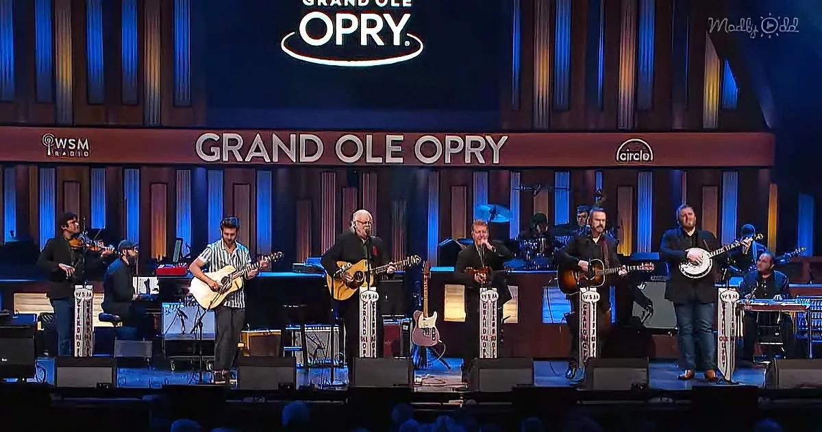 Musicians perform on stage at the Grand Ole Opry with various instruments and a backdrop.