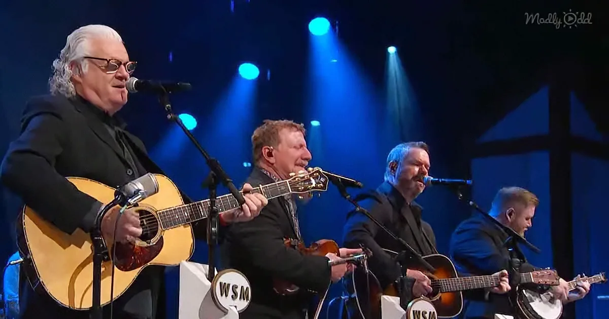An older man plays guitar on stage with a microphone, surrounded by other musicians.