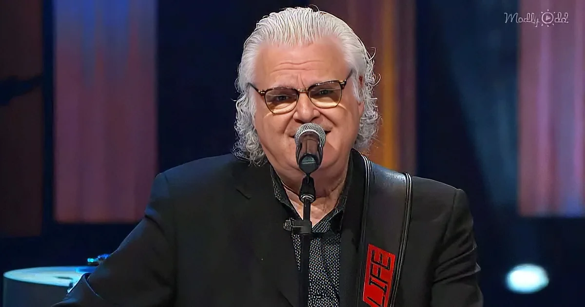 An older man with white hair and glasses sings into a microphone, wearing a black suit with a red logo.